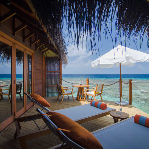 Eine Wasser-Villa-Terrasse mit Blick auf den Ozean, mit Liegestühlen, einem kleinen Tisch, einem Sonnenschirm und einem Schilfdach über der Terrasse.