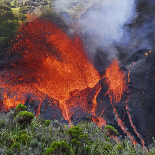 A volcanic eruption with bright red lava spewing out from rocky terrain, emitting smoke and surrounded by green vegetation.