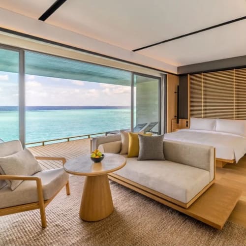 Modern water villa interior with a large glass window offering ocean views, featuring a bed, a small sofa, an armchair, and a round wooden table with a bowl of fruit.