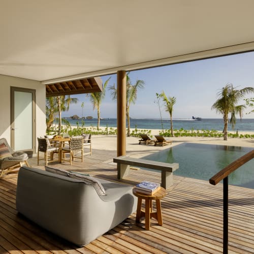A modern outdoor patio featuring wooden flooring, a grey sofa, armchairs, a dining table, and a pool overlooking the beach with palm trees and a boat in the ocean.