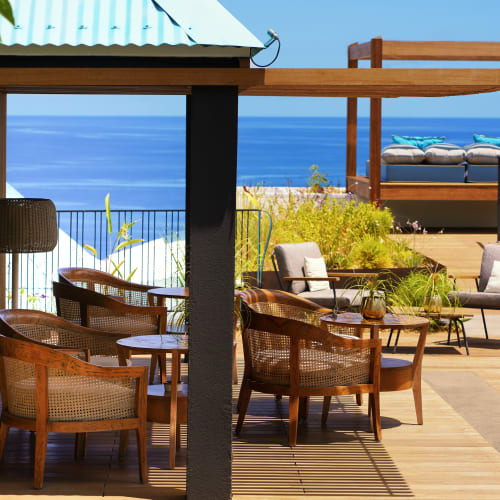 Außenterrasse mit Holzmöbeln, darunter Sessel und Tische unter einem schattigen Dach, mit Blick auf das blaue Meer und eine Tagesliege im Hintergrund auf einem Holzdeck.
