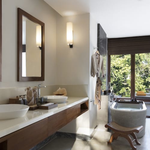 A luxurious bathroom with a double sink vanity, two mirrors, and two wall-mounted lights, leading to an outdoor patio with a relaxing hot tub and a view of greenery.