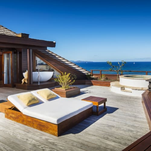 A luxurious rooftop terrace overlooking the ocean with outdoor furniture, a bathtub, and a hot tub, on a sunny day.