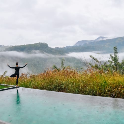 A person practicing yoga on a boardwalk overlooking lush green hills with mist and mountains in the background.