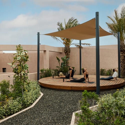 Drei Personen praktizieren Yoga auf einer runden Holzplattform mit Sonnensegel, umgeben von Pflanzen und modernen beigen Gebäuden unter einem leicht bewölkten Himmel.