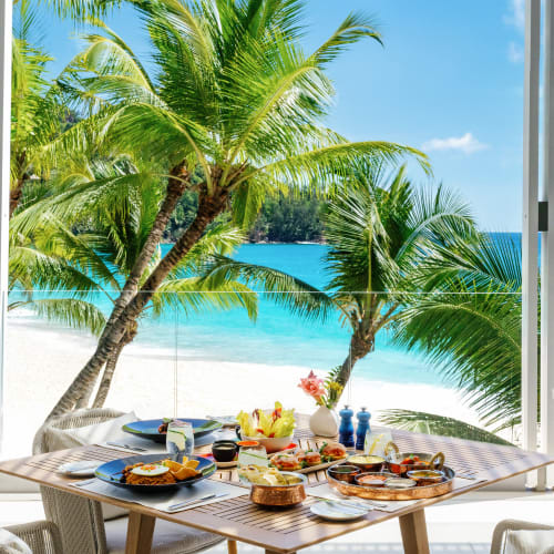A wooden dining table set with various dishes and drinks is placed on a terrace overlooking a tropical beach with turquoise water, white sand, and lush palm trees under a sunny blue sky.