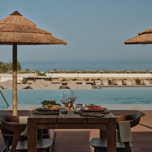 Esstisch mit Tellern, Weingläsern und Essen am Pool unter Strohhschirmen mit Blick auf das Meer im Hintergrund.