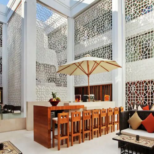 Heller, luftiger Hotel-Lobby mit aufwendigen geometrischen Wandmustern, einem Barbereich mit Holzstühlen und großem Sonnenschirm sowie gemütlicher Sitzecke mit Kissen.