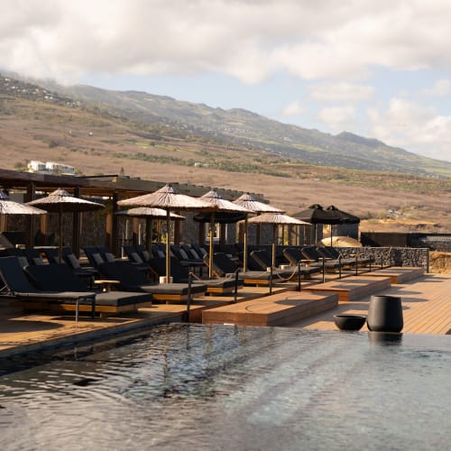 Ein Infinity-Schwimmbecken mit dunklen Liegestühlen und Strohschirmen, die entlang eines Holzdecks angeordnet sind, vor einem Hintergrund aus sanften Hügeln unter einem wolkigen Himmel.
