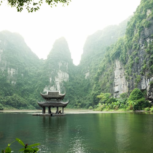 Ein traditionelles Pavillon auf einem ruhigen Fluss, umgeben von hohen grünen Kalksteinbergen in Ninh Binh, Vietnam.