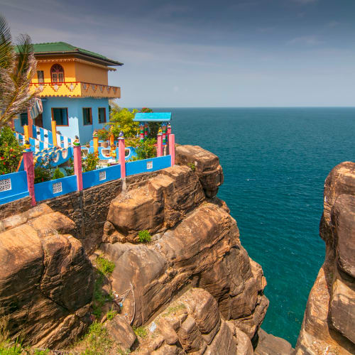 Ein buntes Haus mit blauen Wänden und grünem Dach auf felsigen Klippen mit Blick auf den blauen Ozean unter klarem Himmel.