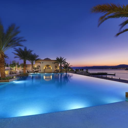 A luxurious hotel pool area with illuminated water and palm trees during sunset or dusk in Aqaba, Jordan.