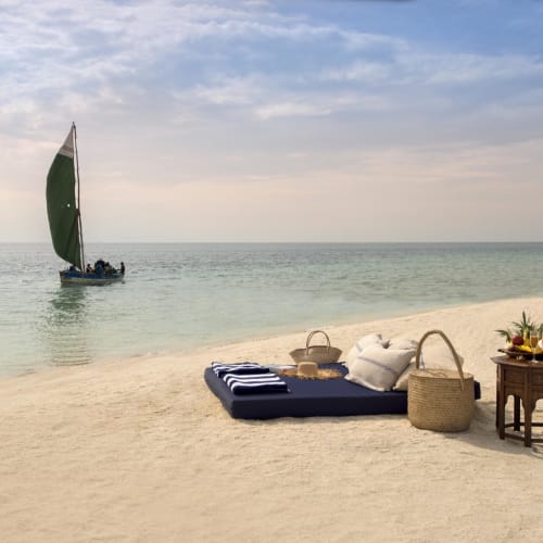 A peaceful beach scene with a sailboat on calm waters and a picnic setup on the sandy shore, including a blanket, pillows, and a small wooden table with snacks and drinks.