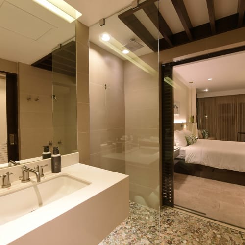 Modern hotel bathroom with white sink, black soap dispensers, and a glass partition leading to a bedroom.