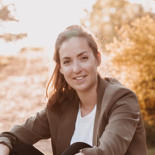 Eine Frau mit braunem Haar, die bei Sonnenuntergang draußen lächelt, trägt eine braune Jacke und ein weißes Oberteil.