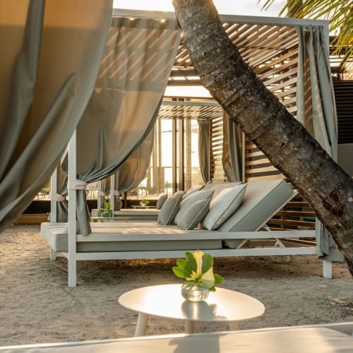 Ein Strand-Loungebereich mit gepolsterten weißen Liegestühlen unter Holzgazebos mit hellgrauen Vorhängen, auf Sand gelegen, mit einer Palme im Vordergrund und sanftem Sonnenlicht, das durchscheint.
