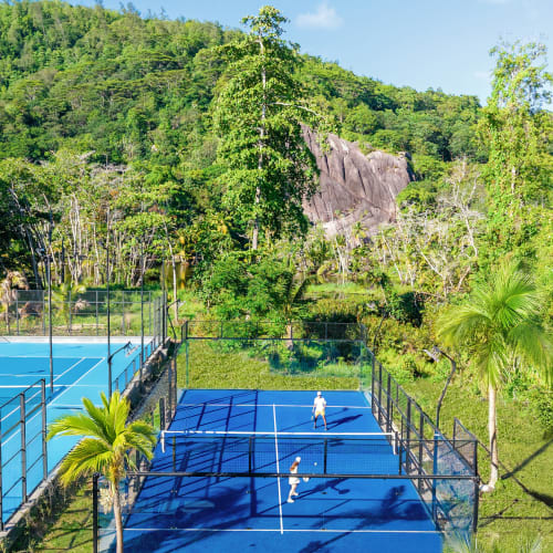 Zwei Personen spielen Padel auf einem blauen Platz, umgeben von Zäunen und Palmen, mit einem bewaldeten Hügel und einem Felsen im Hintergrund unter klarem blauem Himmel.