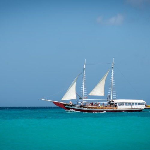 A sailboat with two masts sailing on calm turquoise water under a cloudy sky.