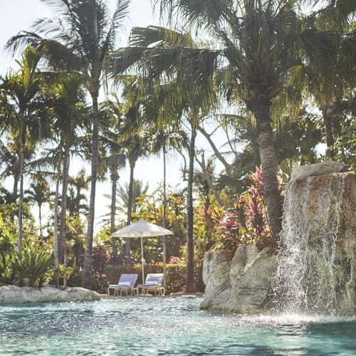 Ein luxuriößer Pool umgeben von tropischen Palmen mit Wasserfall, Liegen am Pool und einem Sonnenschirm in einem tropischen Resort.