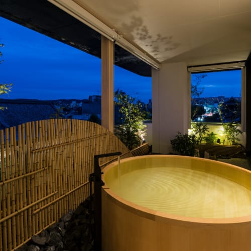 A cozy outdoor hot tub on a balcony at night with city lights and greenery in the background.