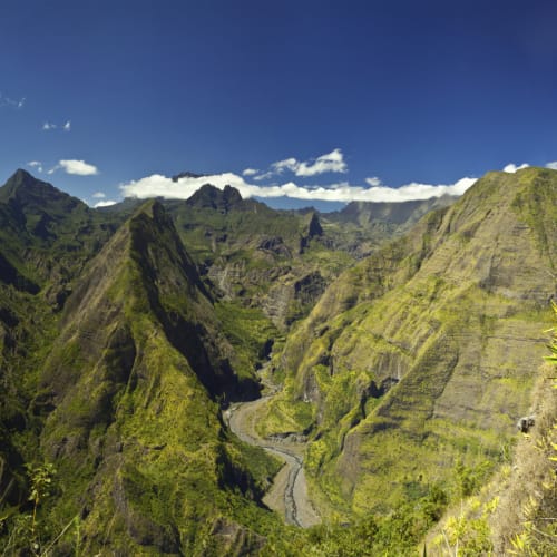Panoramic view of lush green mountains with a winding river valley and some people standing on a lookout point to the right under a partly cloudy blue sky.