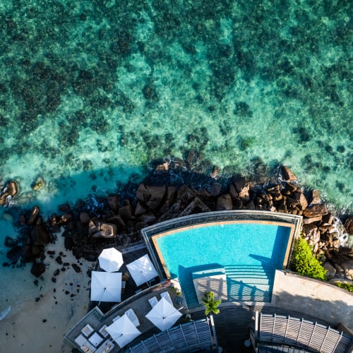Luftaufnahme eines Swimmingpools, der von Sonnenschirmen und Liegestühlen umgeben ist, neben einer felsigen Küste mit klarem türkisfarbenem Wasser.