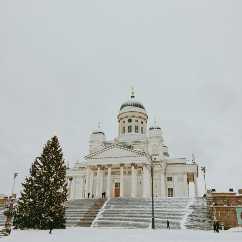 Schneebedeckte Treppen zum Helsinkier Dom im Winter, mit einem großen geschmückten Weihnachtsbaum links und einigen Spaziergängern, unter bedecktem Himmel.