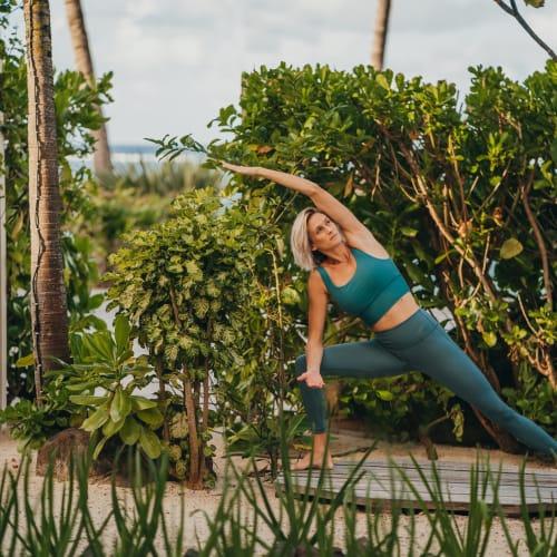 A woman practicing yoga outdoors on a wooden platform surrounded by lush green plants and trees in a tropical setting.