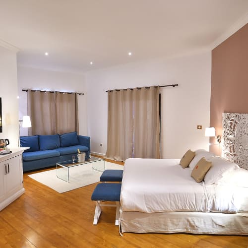 Geräumige Hotelsuite mit einem großen Doppelbett, weiß-beiger Einrichtung, einem blauen Sofa, einem an der Wand montierten Flachbildfernseher und Holzboden.