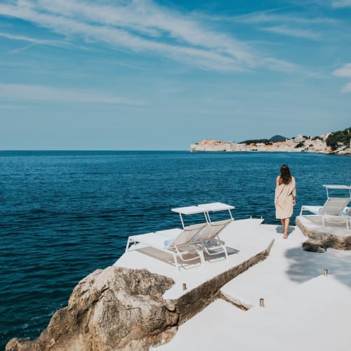 A woman walking along a rocky and white paved coast with sunloungers overlooking the blue sea in Croatia.