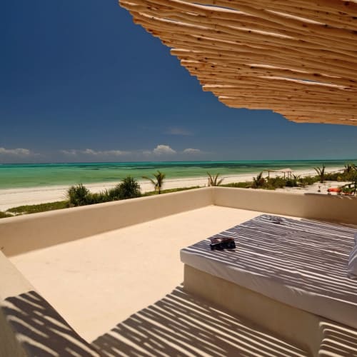 Eine sonnige Terrasse mit Blick auf einen unberührten Strand mit türkisfarbenem Wasser in Zanzibar, mit einem schattigen Lounge-Bereich mit Kissen und einer gestreiften Decke.