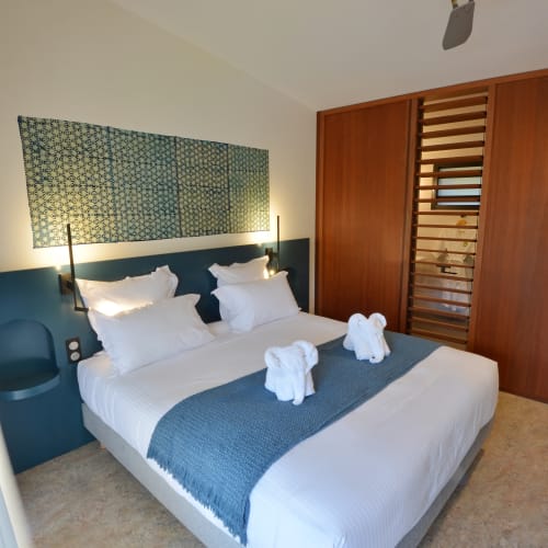 Modernes Schlafzimmer mit großem Doppelbett mit weißer Bettwäsche und blauem Läufer, zwei Handtuch-Elefanten, blauem eingebautem Kopfteil mit seitlichen Regalen und Lampen, einem gemusterten Stoffpaneel über dem Bett, holzverkleideter Wand mit Lamellenabschnitt und Deckenventilator.