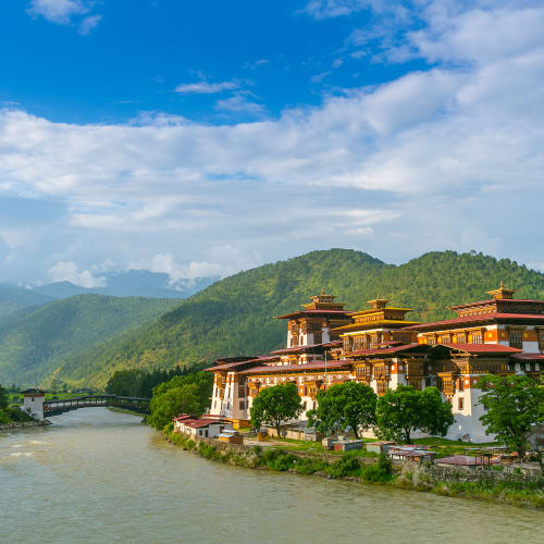 Schönes bhutanisches Kloster mit traditioneller Architektur am Fluss, umgeben von grünen Hügeln unter einem bewölkten Himmel.
