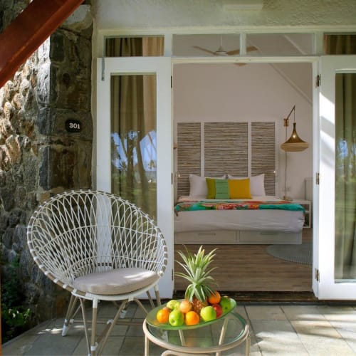 Offene weiße Doppeltüren geben den Blick frei auf ein helles Schlafzimmer mit einem Bett, das mit bunten Kissen und einem naturfasernen Kopfteil dekoriert ist. Draußen auf der Terrasse steht ein weißer Korbsessel und ein runder Glastisch mit einer Schale voller Ananas und verschiedener Zitrusfrüchte.