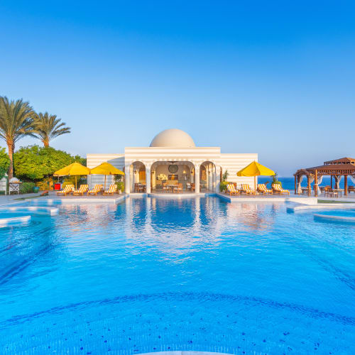Luxury hotel pool area with yellow umbrellas, palm trees, and a domed building in the background, overlooking the sea.