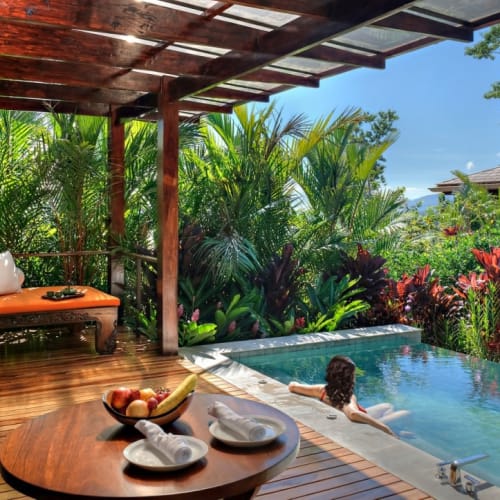 A woman relaxing in a small pool on a wooden terrace surrounded by lush tropical plants.