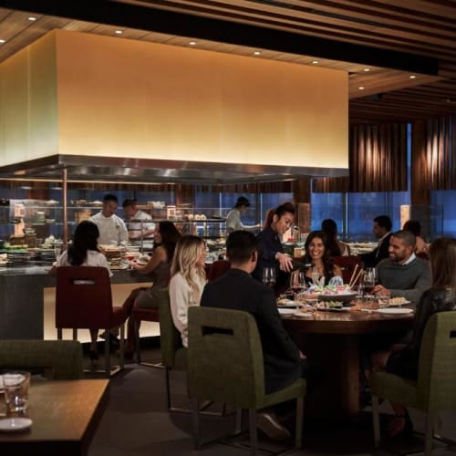 Elegantes Restaurantinterieur mit Gästen, die um einen großen runden Tisch speisen, und Köchen, die hinter der Theke arbeiten.