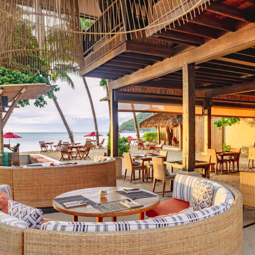 Open-air beachside restaurant with wicker seating, round wooden tables, and tropical decor, overlooking the sea and palm trees under a shaded wooden roof.