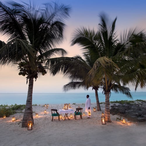 Ein romantisches Privatdinner auf einem Sandstrand mit Palmen, Kerzenlaternen und einer Person in weißer Kleidung mit Blick auf den Ozean bei Sonnenuntergang.