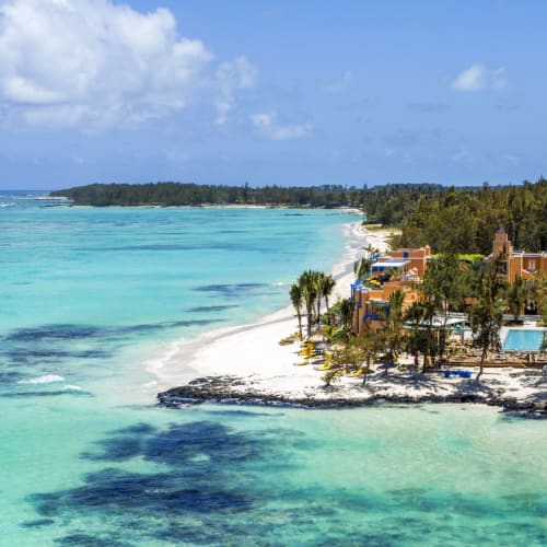 Turquoise sea and white sand beach with a resort nestled among palm trees and greenery under a partly cloudy sky.