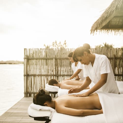 Zwei Spa-Therapeuten geben Kunden eine Massage, die liegend auf Massagetischen im Freien am Wasser sind, mit einem Bambuszaun im Hintergrund.
