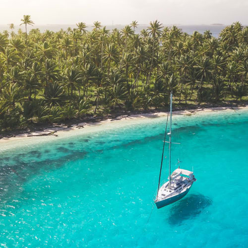 Ein Segelboot treibt nahe einer tropischen Insel mit dichtem Palmenwald und kristallklarem Wasser.