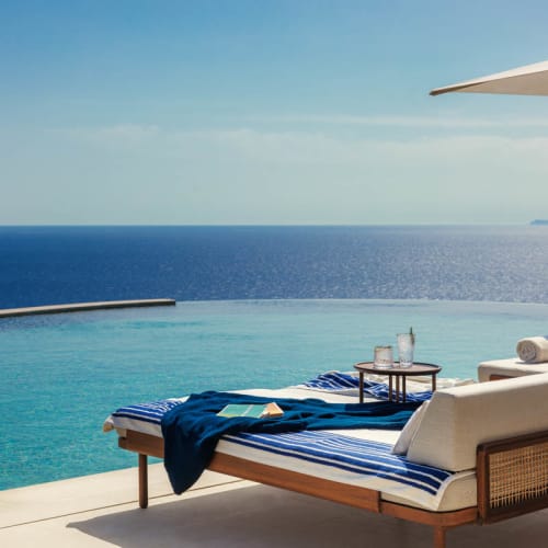 Infinity pool overlooking the sea with sunbeds featuring blue and white striped cushions and a towel, under a large white umbrella on a sunny day.