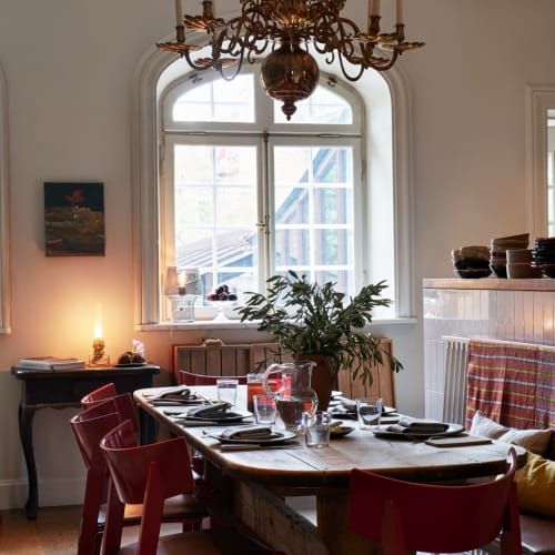 A cozy dining area in a bright kitchen with large arched windows, a wooden table set for a meal with black plates, glasses, and a large green plant as a centerpiece.