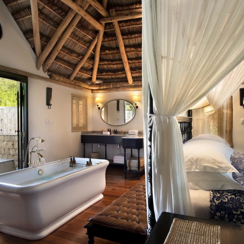 A luxurious interior of a cabana on Benguerra Island with a bathtub near a balcony door and a bed with mosquito netting on the right.