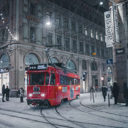 Eine rote Straßenbahn fährt nachts bei Schnee auf Gleisen durch eine Straßenkreuzung mit historischen Gebäuden und Fußgängern in Helsinki.