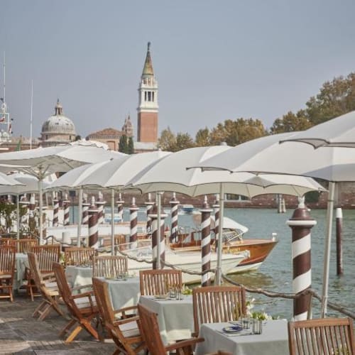 Essterrasse am Wasser mit weißen Sonnenschirmen, Holzgarnituren und Tischen mit weißen Tüchern in Venedig.