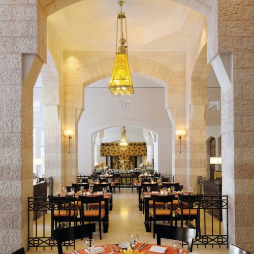 Elegantes Restaurant-Innenraum mit hohen Steinbögen, hängenden goldenen Kronleuchtern und sorgfältig gedeckten Tischen mit orangefarbenen Tischdecken und Blumenarrangements.