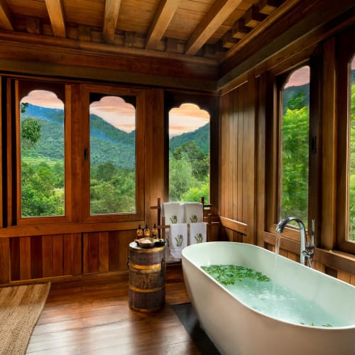 A luxurious wooden bathroom with a freestanding bathtub overlooking lush green mountains through large windows.