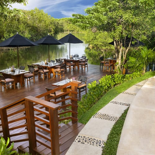 A restaurant terrace built on a wooden deck surrounded by lush greenery and overlooking a river, with tables set up under black umbrellas.